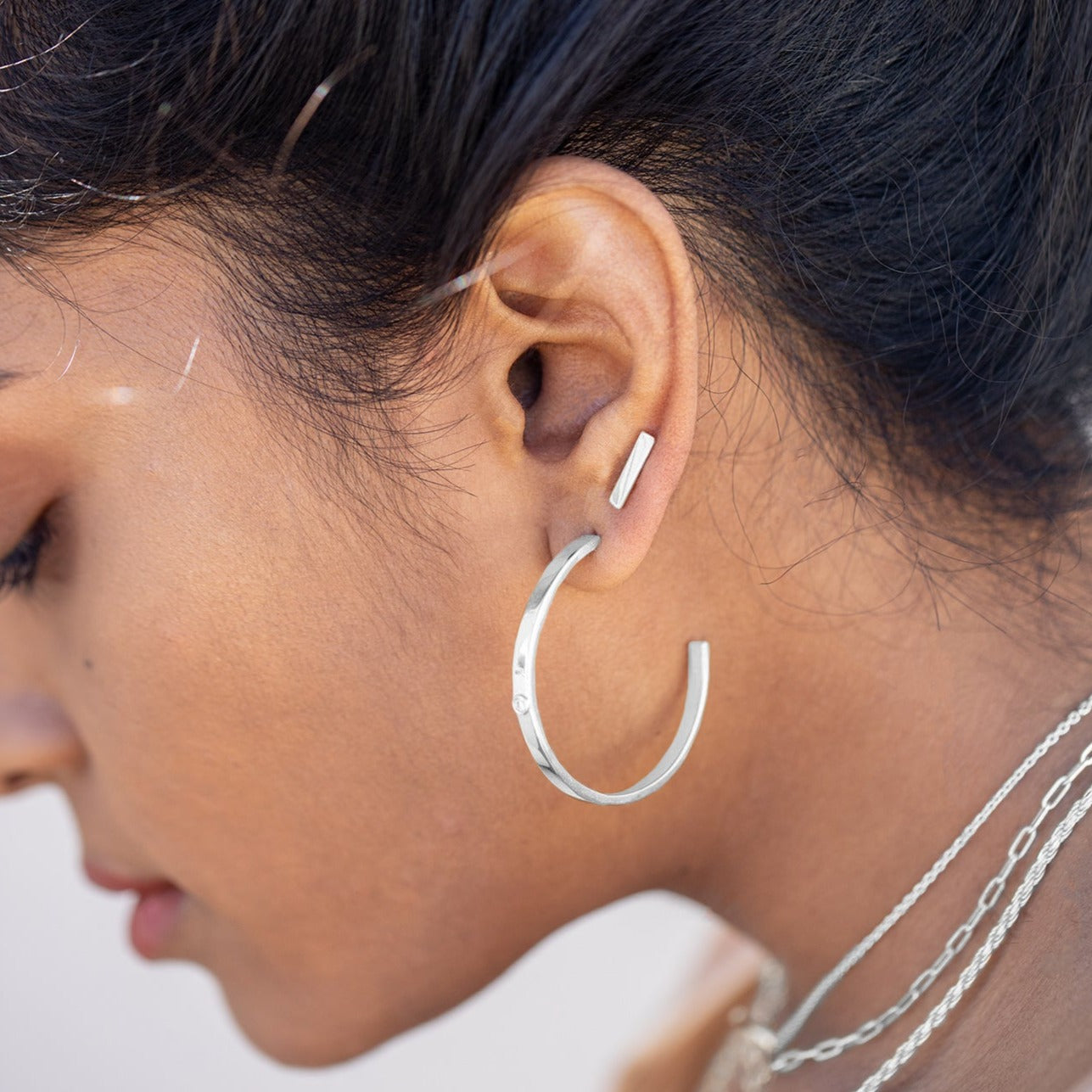 Sterling silver stud earrings positioned in the second piercing of a model's ear. Large hoop earrings with lab grown diamonds are in the first position. 