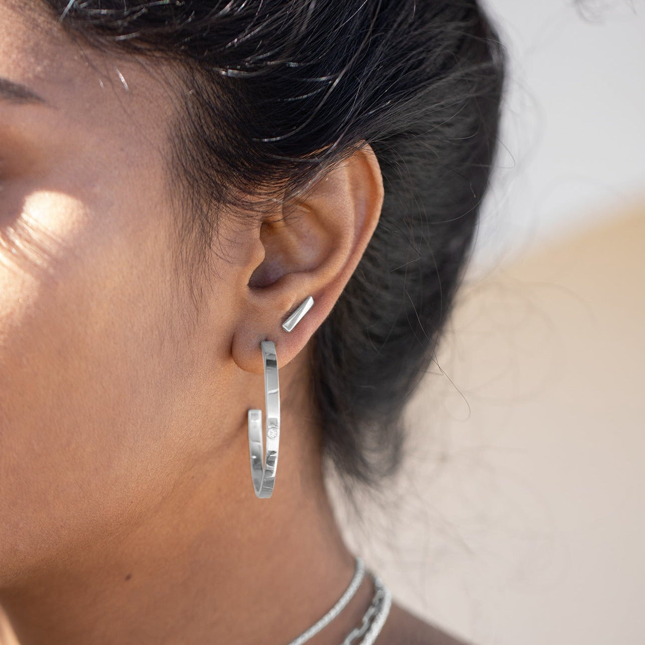Sterling silver stud earrings positioned in the second piercing of a model's ear. Large hoop earrings with lab grown diamonds are in the first position. 