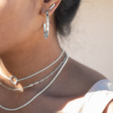 A close up of a model wearing three silver necklaces and a silver hoop earring with a silver stud earring in the second piercing. 