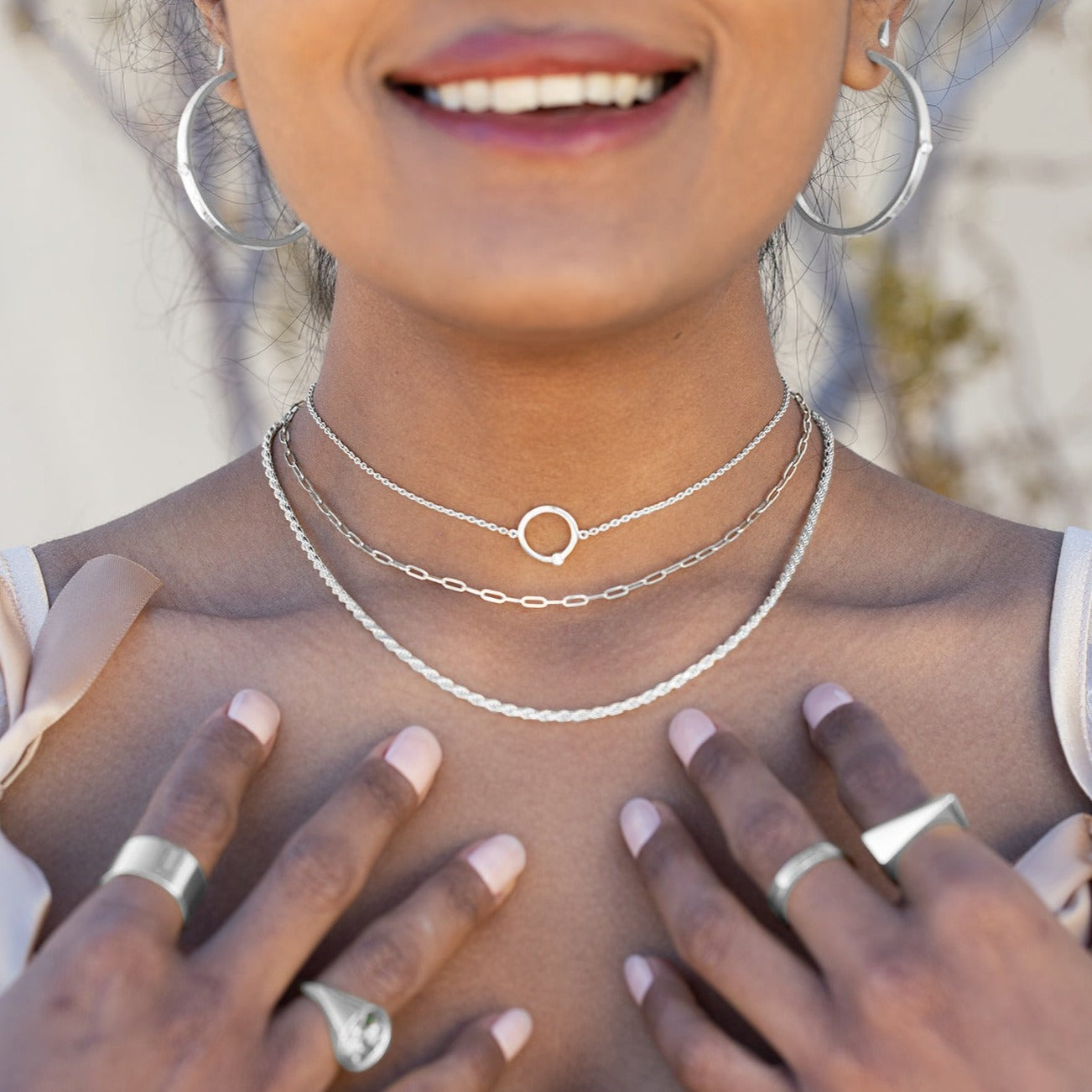 A model wearing three silver necklaces, silver rings, and silver hoop earrings with her hands on her chest and greenery and a white wall behind her. 