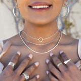 A model wearing three silver necklaces, silver rings, and silver hoop earrings with her hands on her chest and greenery and a white wall behind her. 