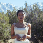 A model wearing gold jewelry outside, holding a small travel case in a satin dress outside. She is outside with bushes and mountains behind her.