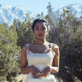 A model wearing gold jewelry outside, holding a small travel case in a satin dress outside. She is outside with bushes and mountains behind her.
