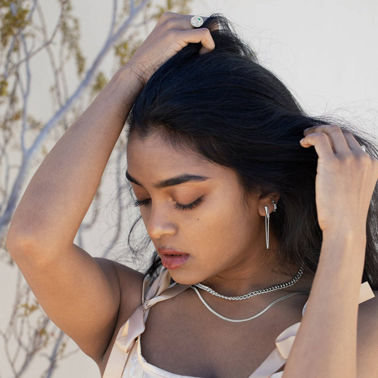 A model running her hands through her hair wearing silver jewelry. She is wearing a pink satin dress and there is greenery and a white wall in the background. 