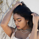 A model running her hands through her hair wearing silver jewelry. She is wearing a pink satin dress and there is greenery and a white wall in the background. 