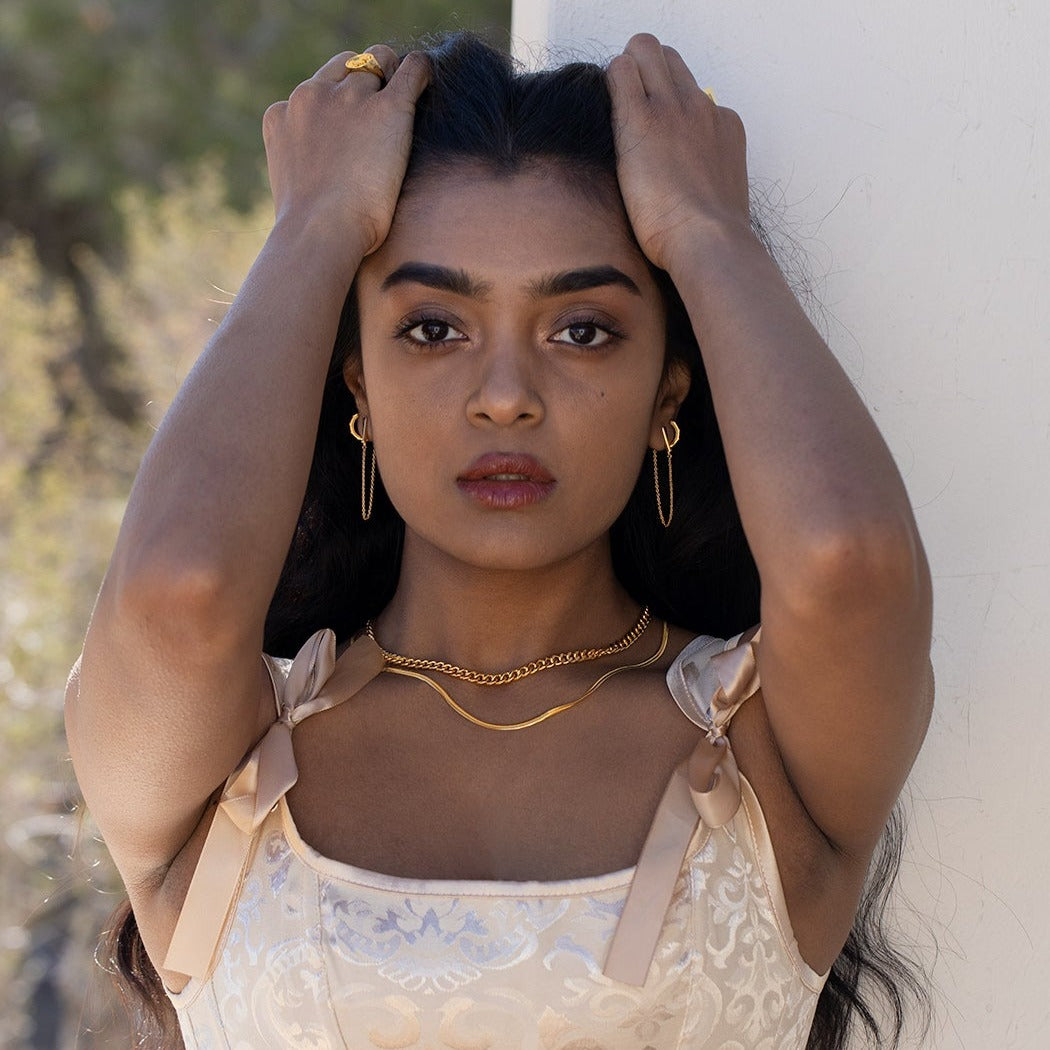 A model running her hands through her hair, wearing gold vermeil jewelry and a pink satin dress with bows on the straps with a white wall and greenery in the background. 