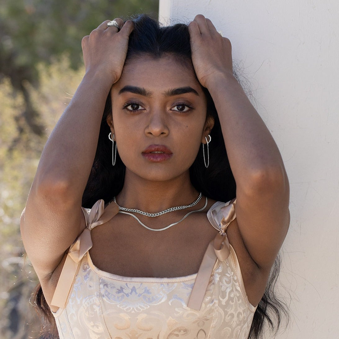 A model running her hands through her hair wearing silver jewelry. She is wearing a pink satin dress and there is greenery and a white wall in the background. 