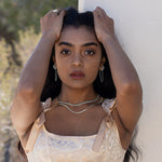 A model running her hands through her hair wearing silver jewelry and a pink satin dress with bows on the straps with greenery and a white wall in the background. 
