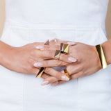 Gold and rose gold toned chunky rings on a model's intertwined fingers. She is wearing a white dress in a studio. 