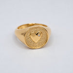 A gold signet ring featuring a textured oval face with a polished heart motif and a lab-grown diamond accent stone, photographed on a clean light gray background.