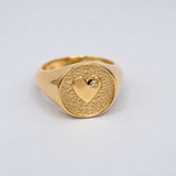 A gold signet ring featuring a textured oval face with a polished heart motif and a lab-grown diamond accent stone, photographed on a clean light gray background.