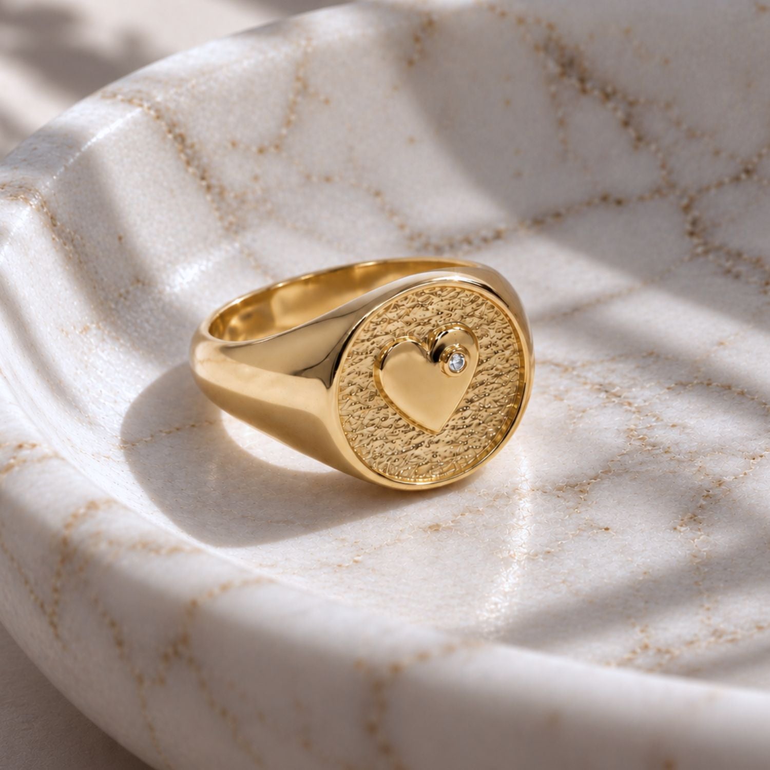 A gold signet ring with a textured face and heart with lab-grown diamond, resting on a white marble tray, with directional sunlight.