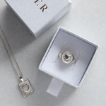 A sterling silver pendant necklace featuring a square textured box with a heart-shaped cutout in the center, suspended from a delicate silver chain against a plain white background with a silver signet ring in a Cier box.