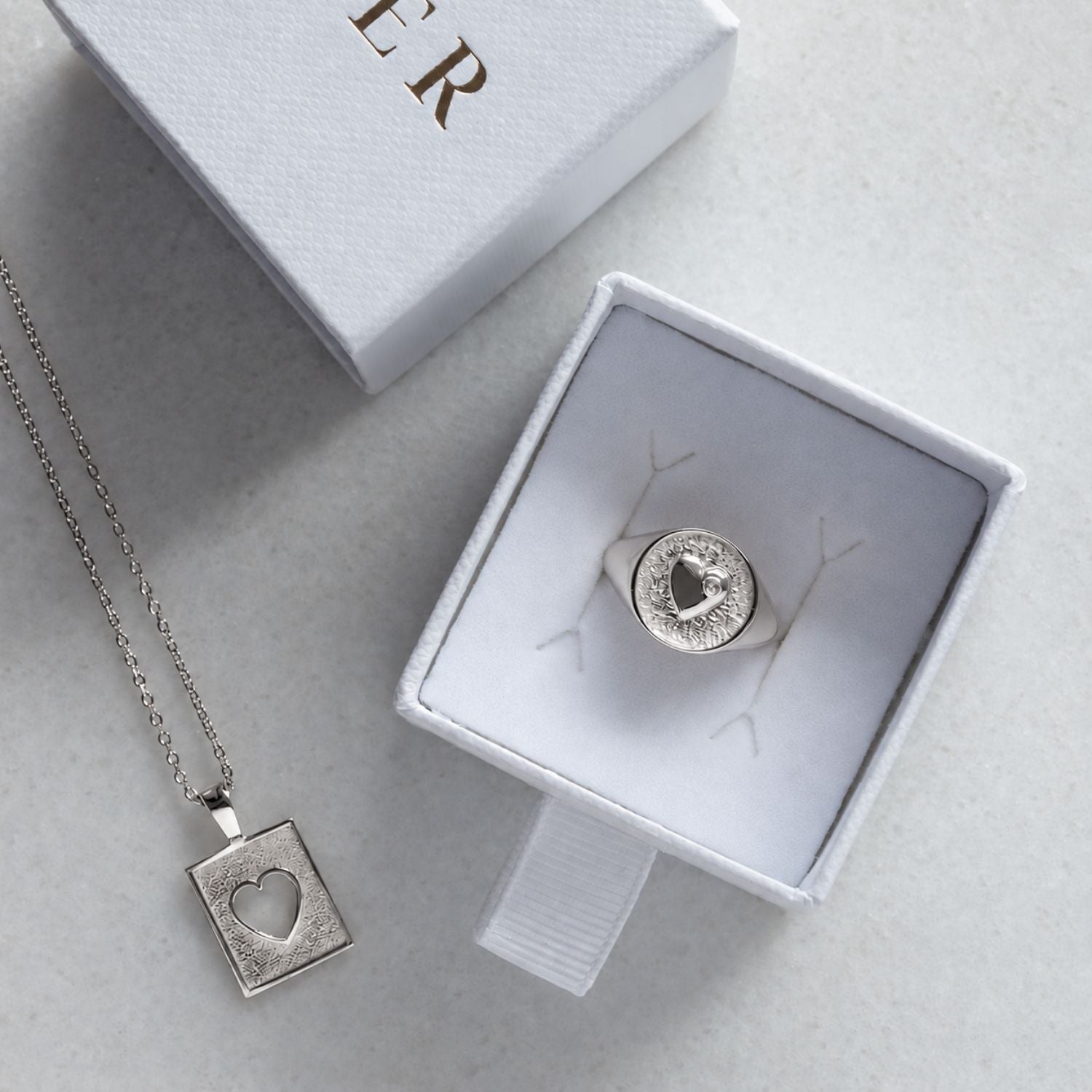 A sterling silver pendant necklace featuring a square textured box with a heart-shaped cutout in the center, suspended from a delicate silver chain against a plain white background with a silver signet ring in a Cier box.