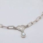 A silver paperclip chain necklace featuring a polished heart-shaped pendant and a small teardrop-shaped white pearl charm, shown on a clean light gray background.
