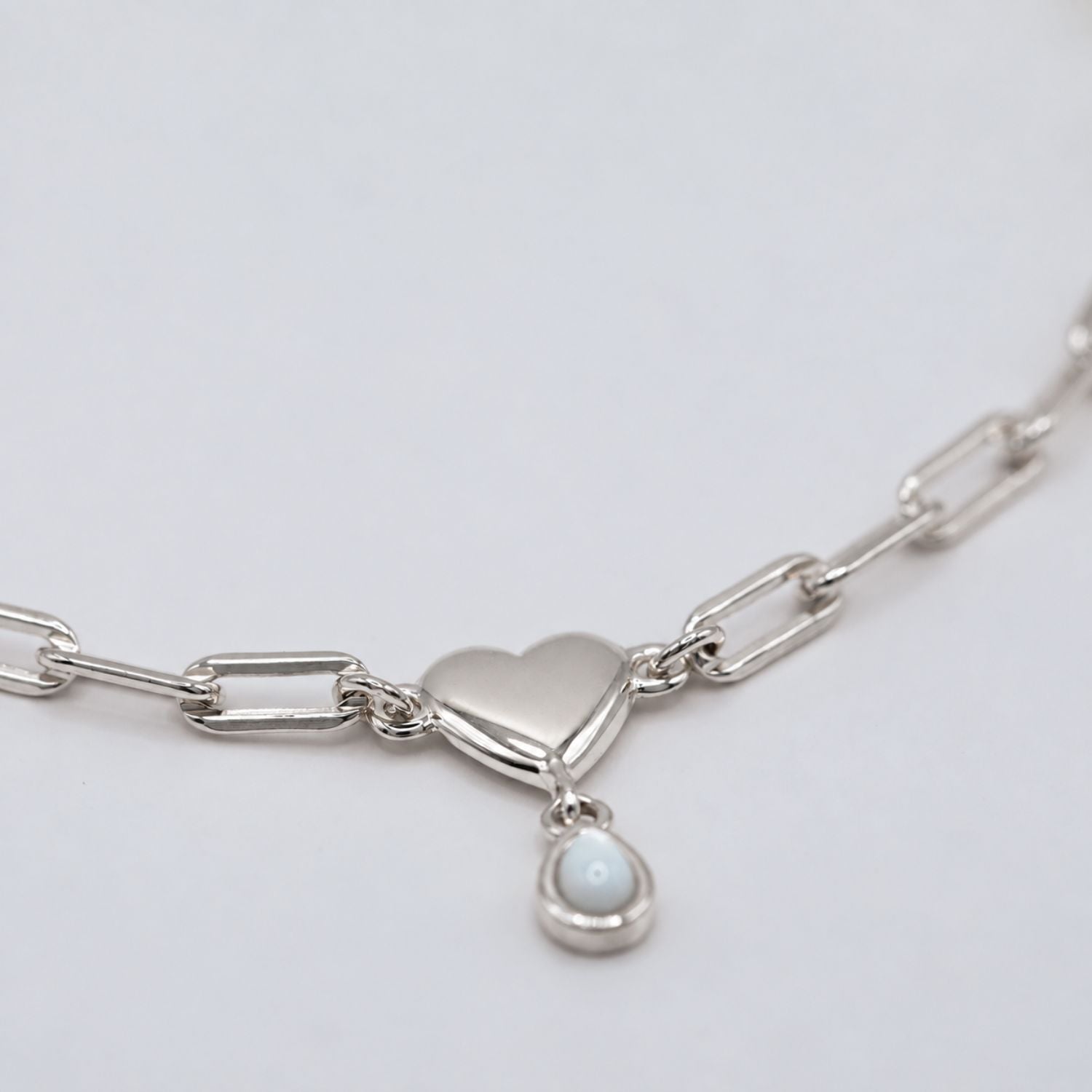 A silver paperclip chain necklace featuring a polished heart-shaped pendant and a small teardrop-shaped white pearl charm, shown on a clean light gray background.