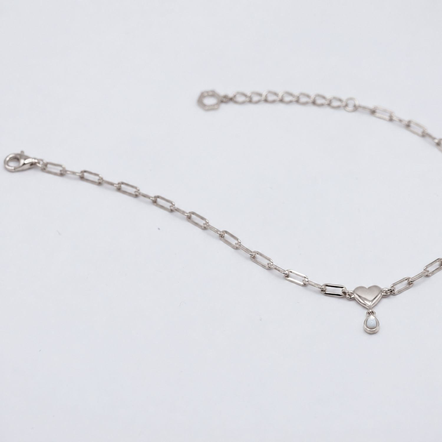 A silver paperclip chain bracelet with a small heart charm and teardrop pearl, displayed on a clean white background.