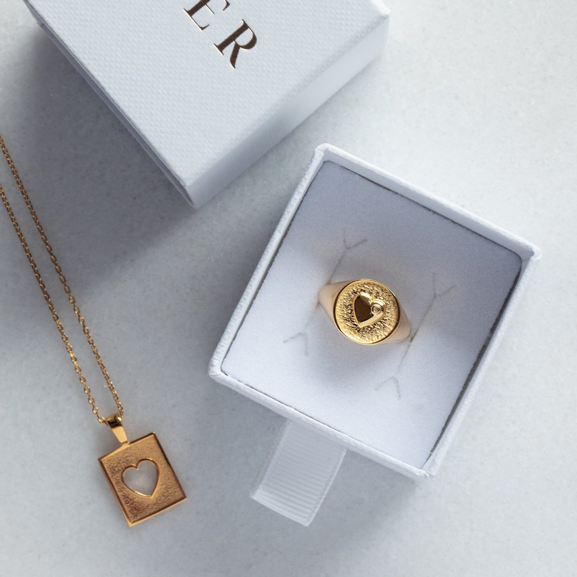 A gold pendant necklace featuring a square textured box with a heart-shaped cutout in the center, suspended from a delicate gold chain against a plain white background with a gold signet ring in a Cier box.