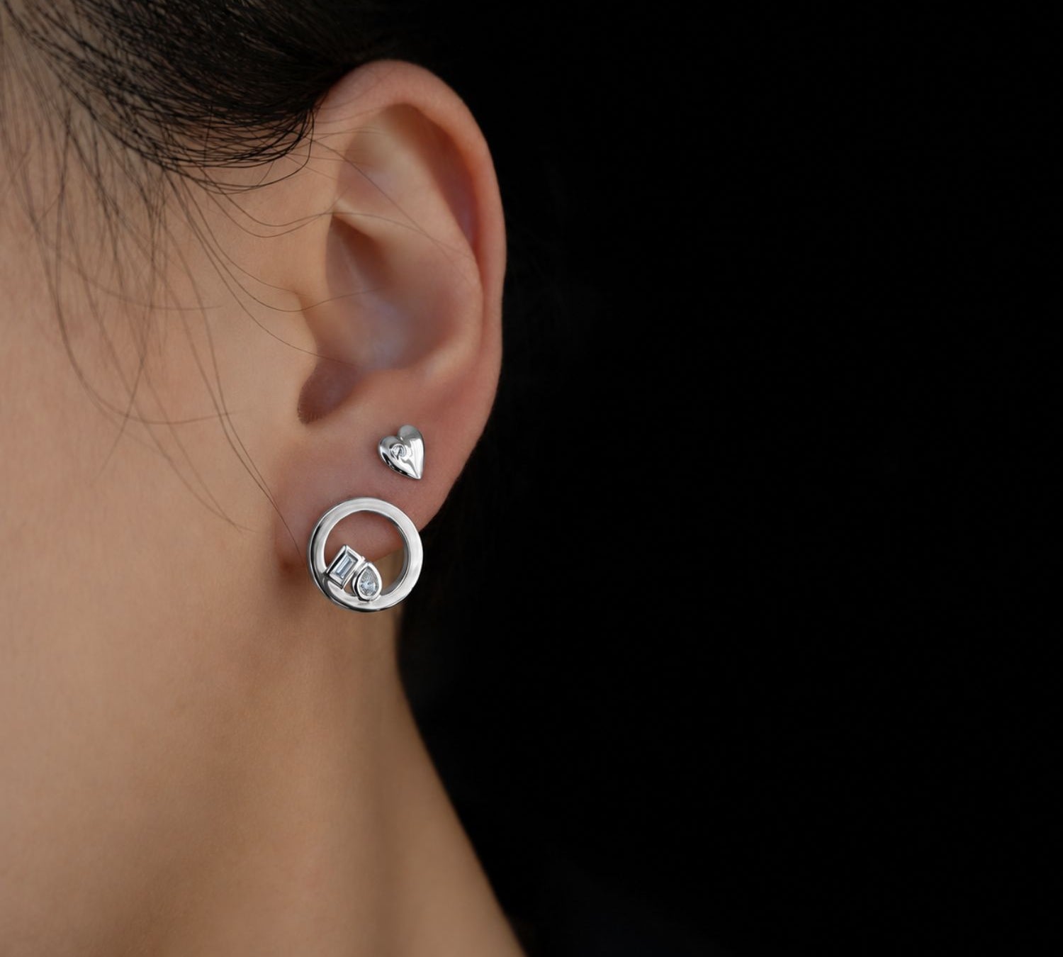 A close-up side profile of a person's ear wearing a silver hoop earring with a small heart-shaped stud and a toi et moi front facing hoop earring, set against a solid black background.