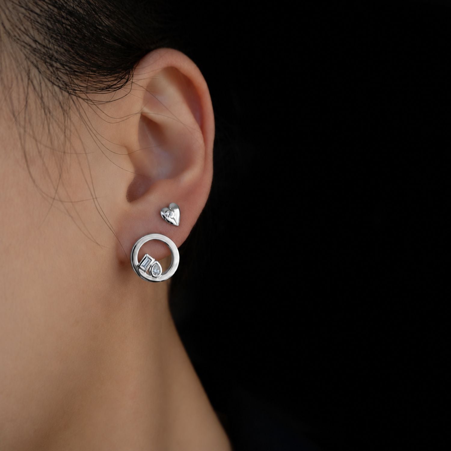 A close-up side profile of a person's ear wearing a silver hoop earring with a small heart-shaped stud and a toi et moi front facing hoop earring, set against a solid black background.