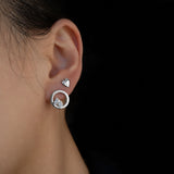 A close-up side profile of a person's ear wearing a silver hoop earring with a small heart-shaped stud and a toi et moi front facing hoop earring, set against a solid black background.