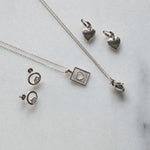 Silver jewelry set including a heart pendant necklace, a square heart pendant necklace, a pair of heart-shaped earrings, and a pair of hoop earrings with lab grown diamonds, arranged on a light marble background.