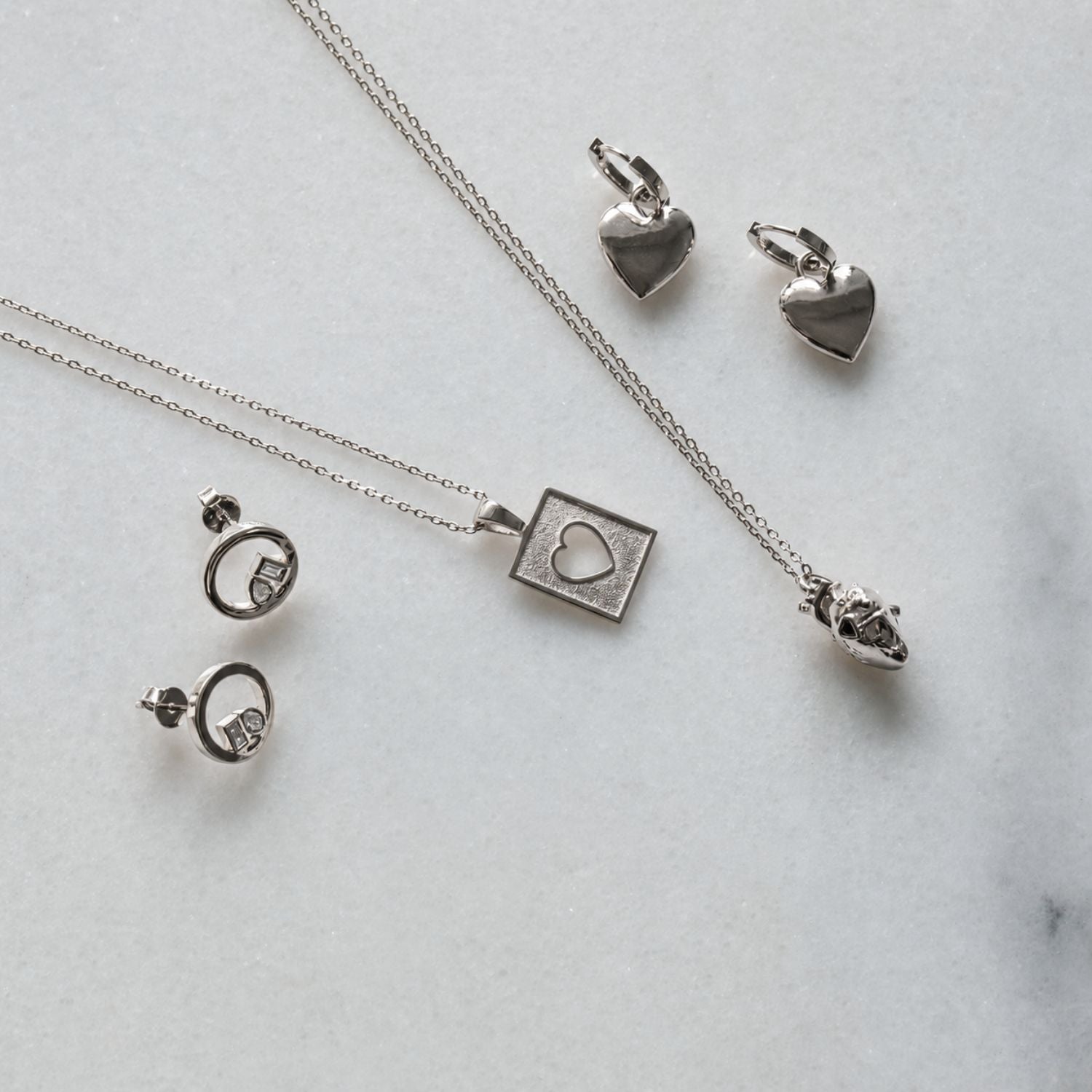 Silver jewelry set including a heart pendant necklace, a square heart pendant necklace, a pair of heart-shaped earrings, and a pair of hoop earrings with lab grown diamonds, arranged on a light marble background.