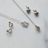Silver jewelry set including a heart pendant necklace, a square heart pendant necklace, a pair of heart-shaped earrings, and a pair of hoop earrings with lab grown diamonds, arranged on a light marble background.