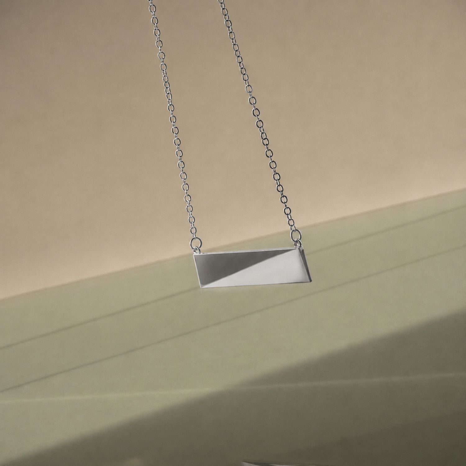 A silver necklace on a delicate chain with a green and grey background and reflection on the bottom of the image. 
