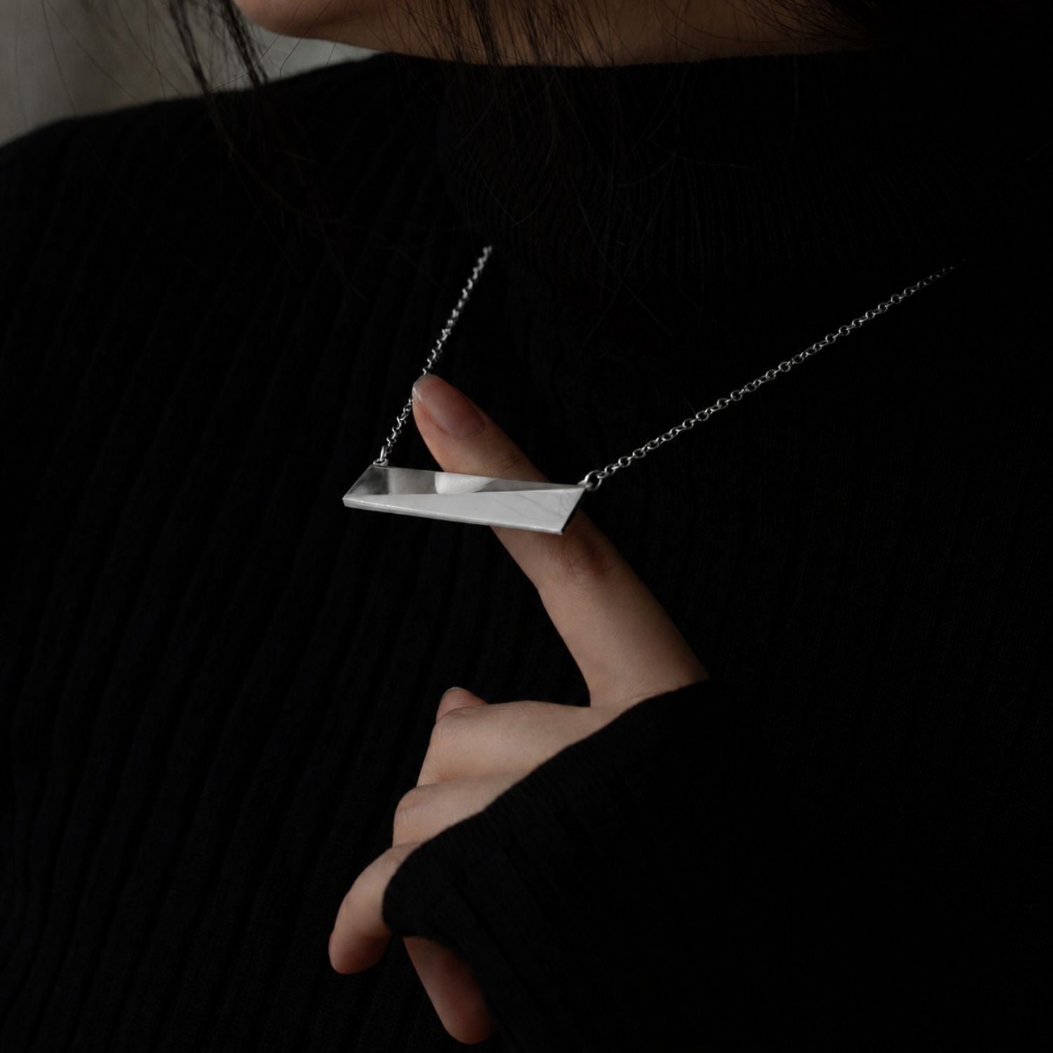 A sterling silver bar necklace on a model with her pointer finger bringing it away from her chest. She is wearing a black turtle neck.