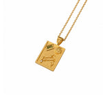 A gold pendant necklace featuring a rectangular design with astrological symbols, including the Libra zodiac sign, a sun motif, and small green and clear gemstones, shown on a white background.
