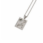 A silver-toned rectangular pendant necklace featuring an engraved zodiac symbol, a flower motif, and a small green gemstone, shown on a clean white background.