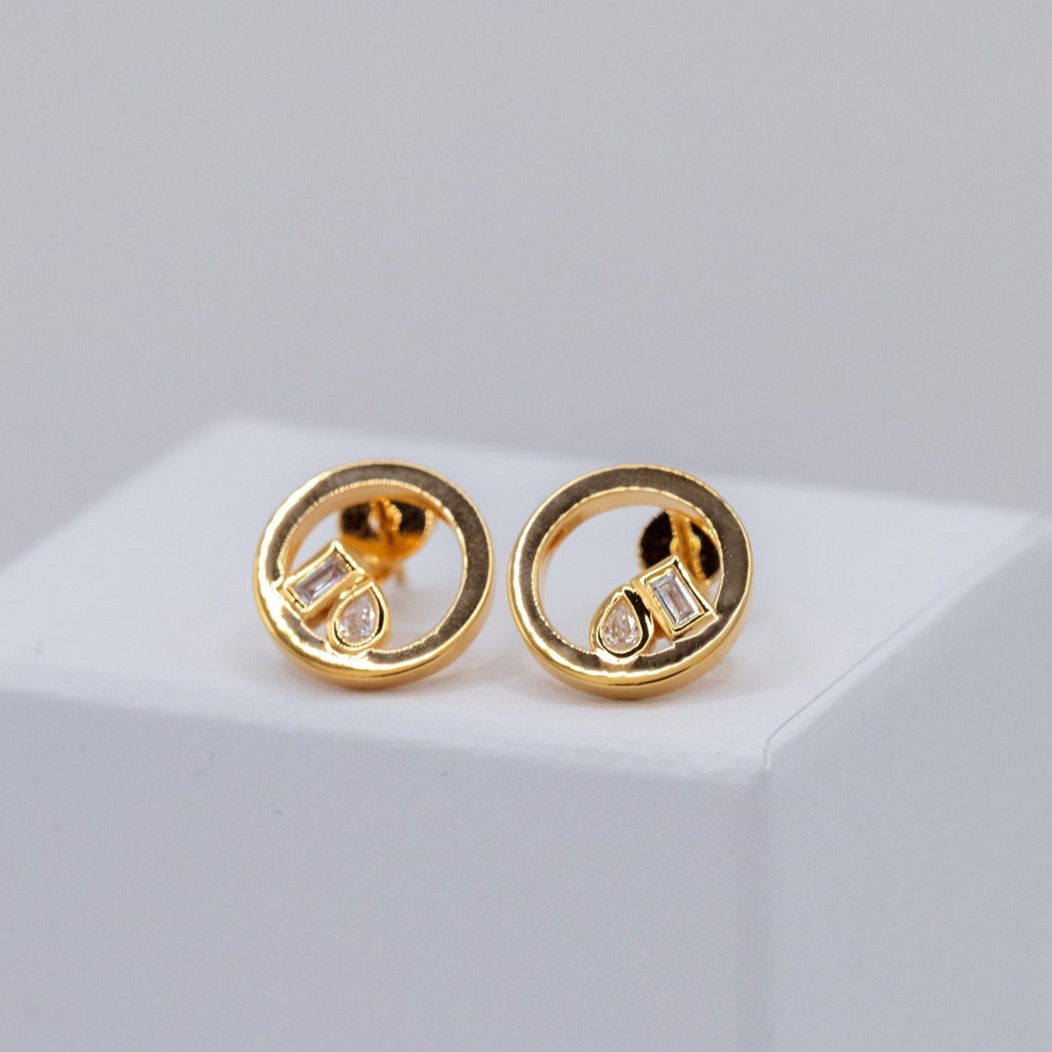 A pair of gold circular stud earrings with baguette and pear-cut lab-grown diamonds, shown on a clean white background.
