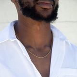A man with an open white button up and a gold rope chain around his neck. A white wall is in the background. 