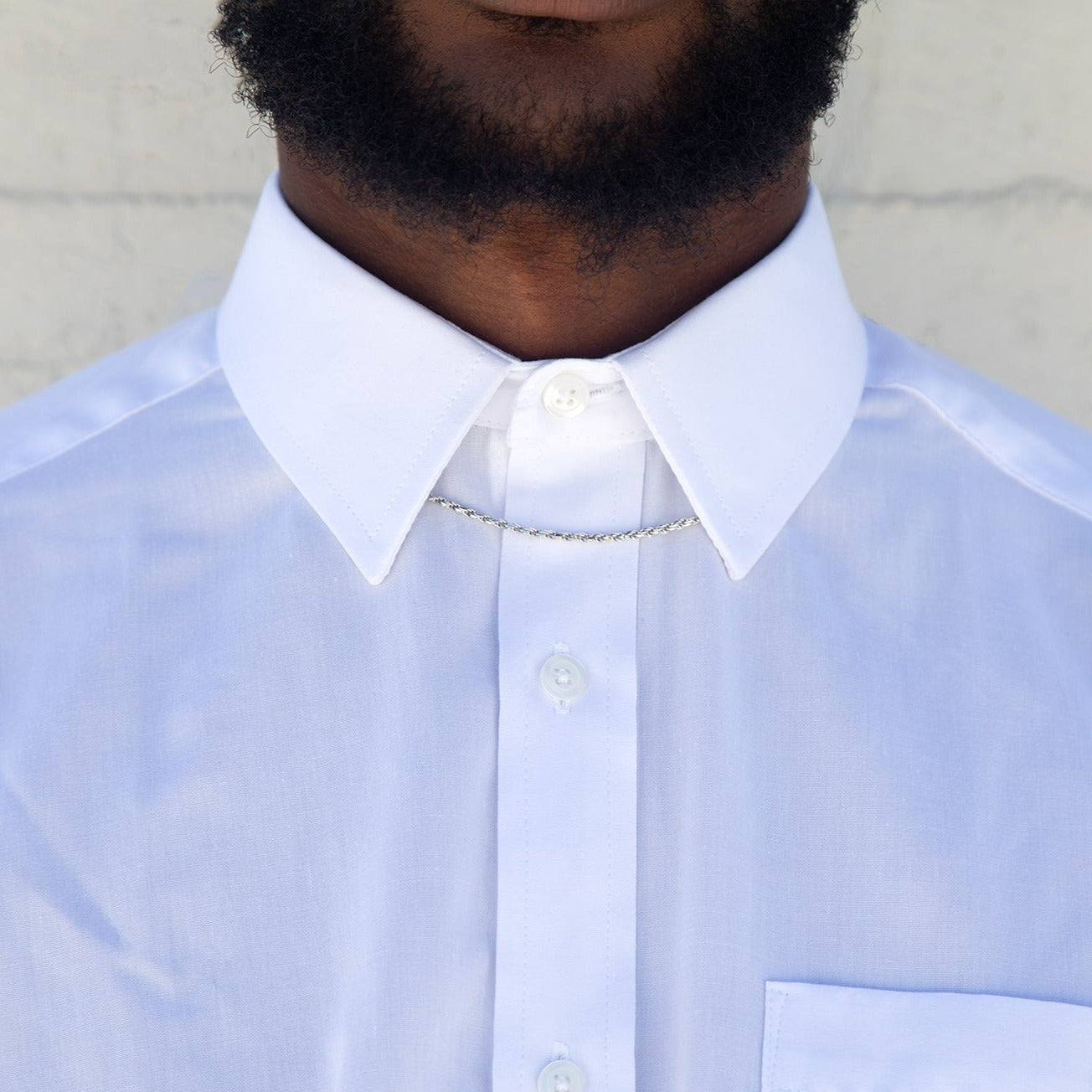 A man wearing a sterling silver rope chain under the collar of a white button up shirt with a white wall behind him. 