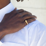 A man's hand on his shoulder with a gold ring on his pinky finger. He's wearing a white shirt and the background is off-white. 