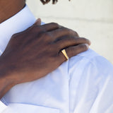 A man's hand on his shoulder with a gold ring on his pinky finger. He's wearing a white shirt and the background is off-white. 