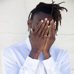 A man wearing a ring on each of his pinky fingers. He is covering his face with hands. Wearing a white shirt. He has a hoop earring and there is a white background.