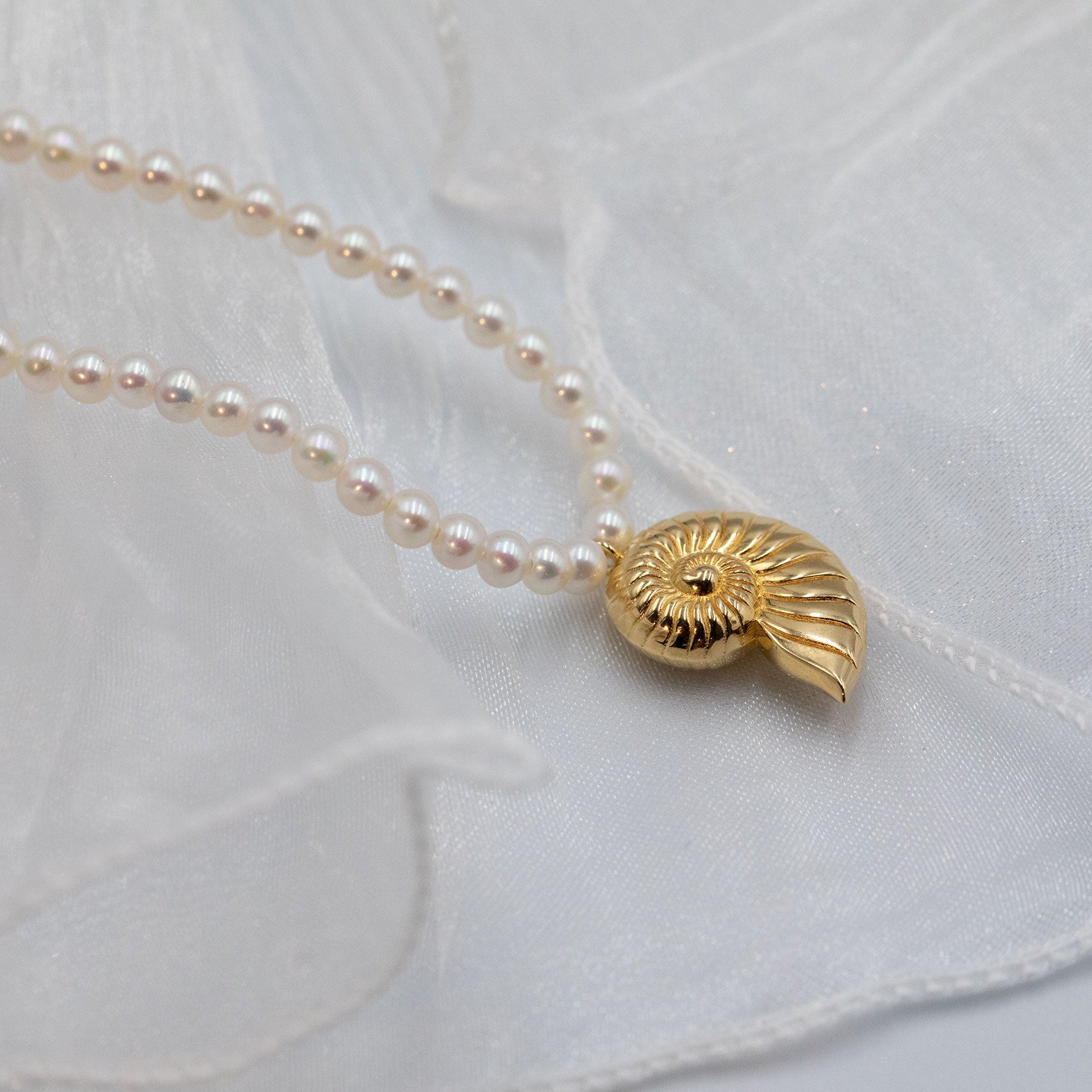 A delicate necklace with small white pearls and a gold shell-shaped pendant, laid on soft white fabric.