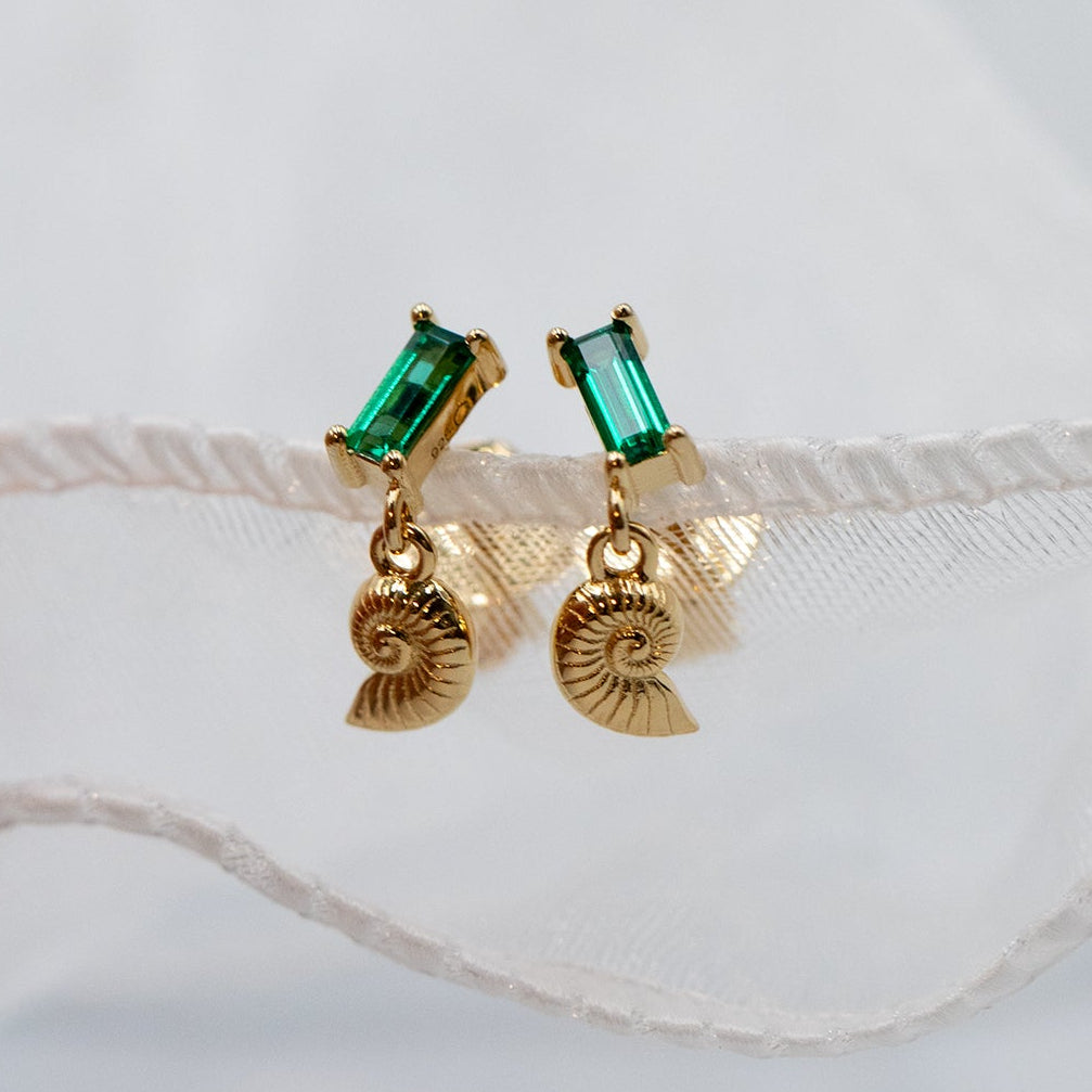 A pair of gold vermeil dangle earrings featuring rectangular green emerald-cut crystals on the studs and a matching gold nautilus shell charm hanging below, displayed against a soft white fabric background.
