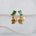 A pair of gold vermeil dangle earrings featuring rectangular green emerald-cut crystals on the studs and a matching gold nautilus shell charm hanging below, displayed against a soft white fabric background.