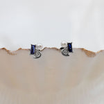 A pair of silver stud earrings with rectangular blue gemstones and small white pearl accents, displayed on the edge of a beige clam shell against a white background.