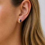 A close-up of a woman's ear wearing a small gold spiral stud earring with a dark blue rectangular stone and a tiny white pearl accent.