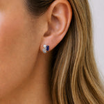 A close-up of a woman's ear wearing a small silver stud earring with a dark blue rectangular stone and a white pearl accent.