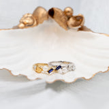 Two rings displayed on a white clam shell: one yellow gold ring with a pear-cut sapphire and round pearls, and one silver band with clear stones, set against a soft white draped fabric background.