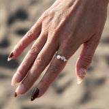 A close-up of a person's hand resting against a sandy background, wearing a silver ring with a pearl and a small blue accent on the ring finger.