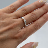 A close-up of a hand wearing a silver ring with a central white pearl and small clear stones on the ring, set against a soft light gray background.