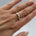 A close-up of a hand wearing a silver ring with a central white pearl and small clear stones on the ring, set against a soft light gray background.