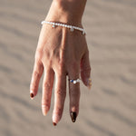 A delicate pearl beaded bracelet with small silver charms, on a model's wrist with sand background.