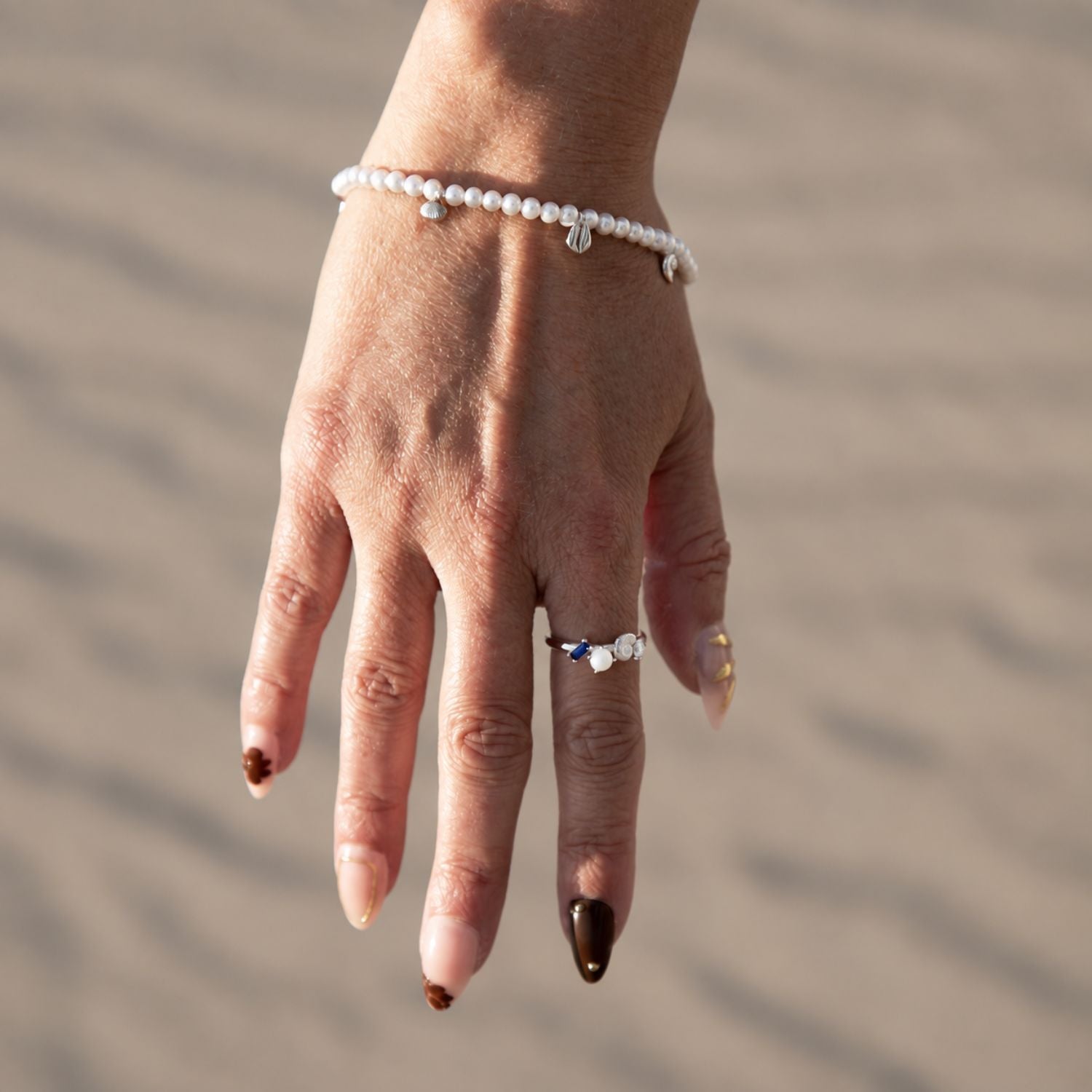A delicate pearl beaded bracelet with small silver charms, on a model's wrist with sand background.